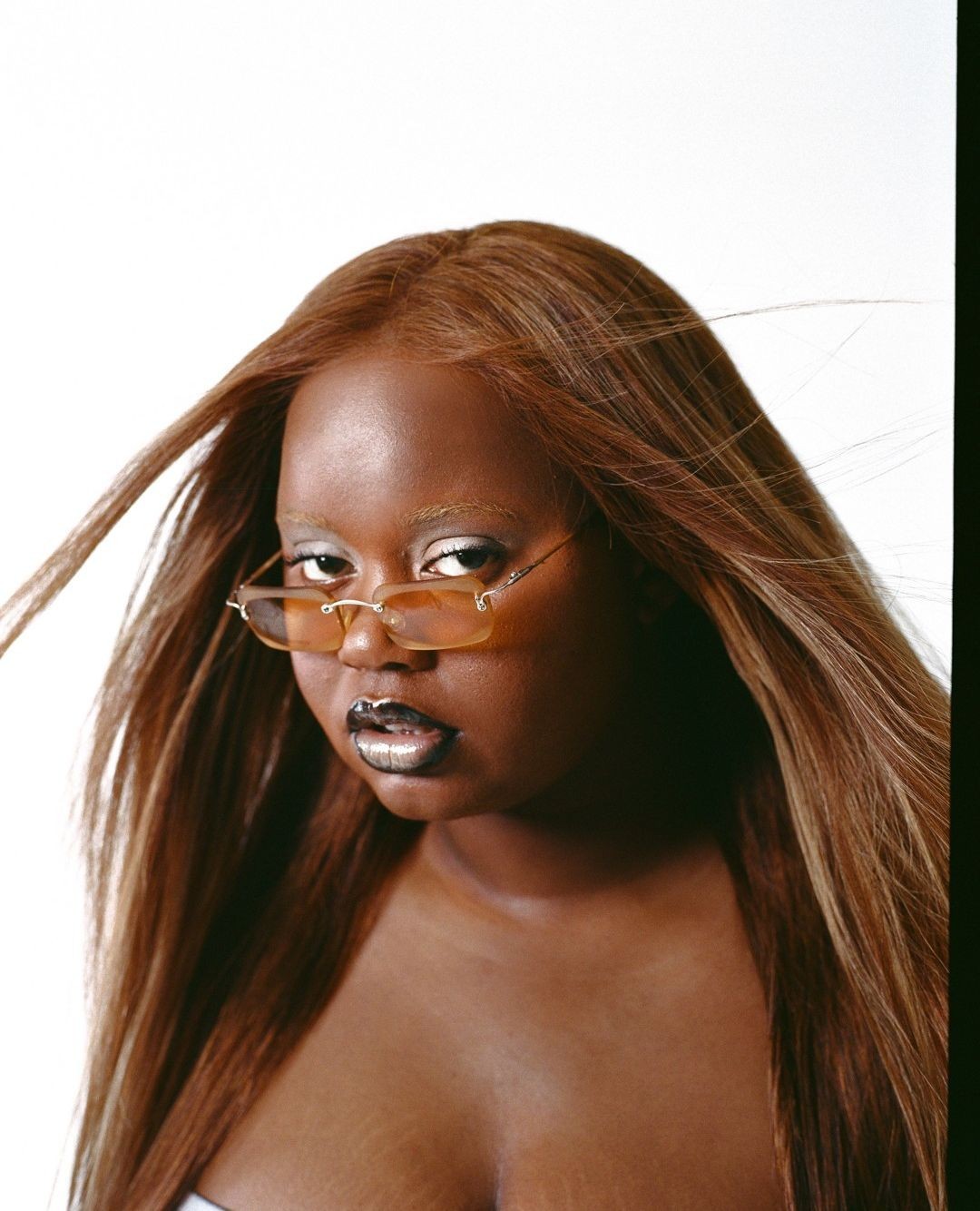 Close-up portrait of a dark-skinned woman with long, caramel-colored hair, wearing peach sunglasses, and silver lipstick against a white backdrop.