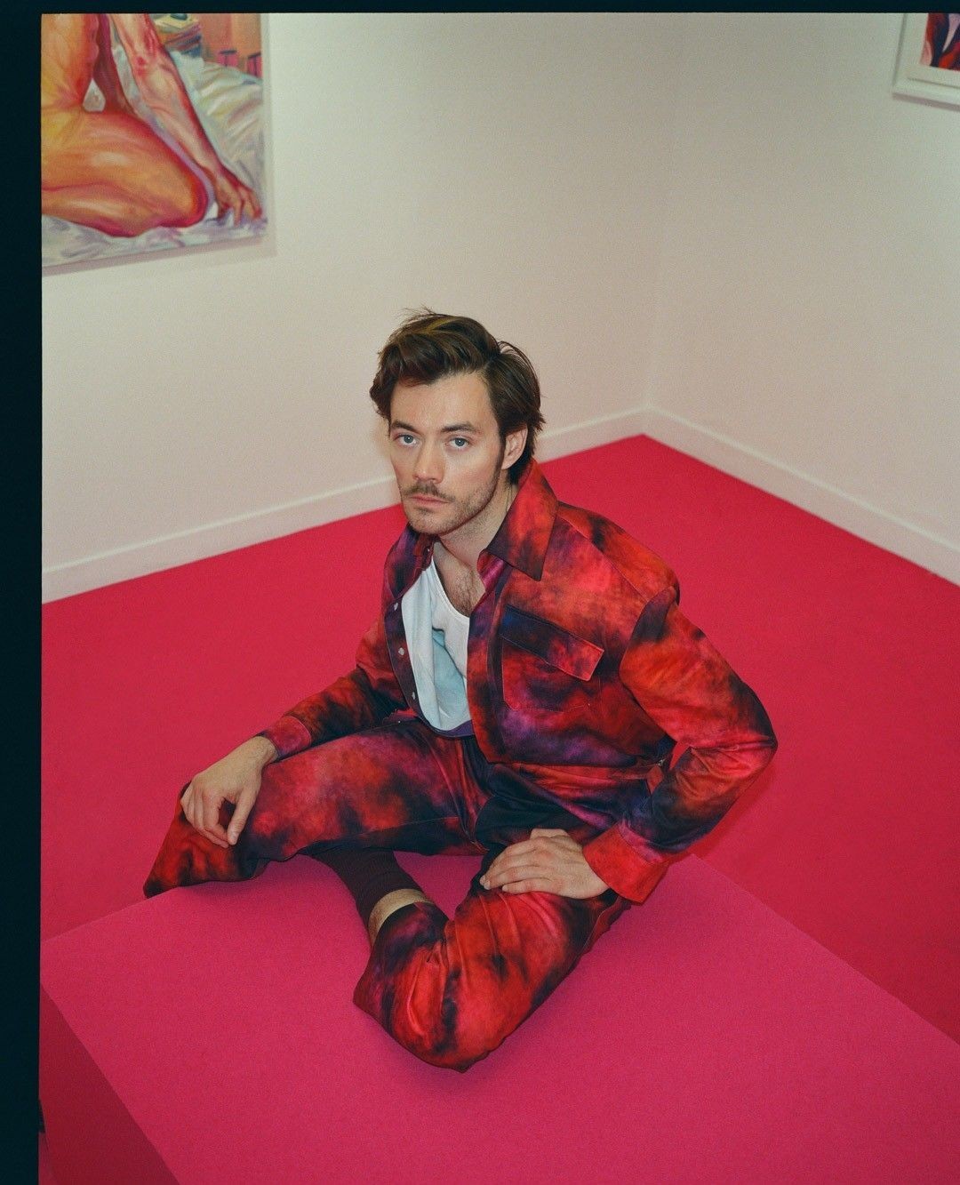 A brunette man sits on a pink cube in a room with white walls, wearing a red and blue patterned suit and white t-shirt with socks.