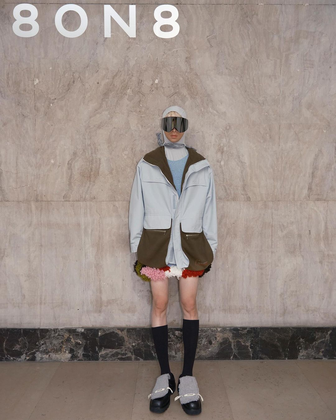Fashion model poses in light blue jacket with olive green pockets, colorful pompom skirt, black socks and shoes, with goggles.