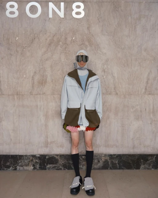 Fashion model poses in light blue jacket with olive green pockets, colorful pompom skirt, black socks and shoes, with goggles.