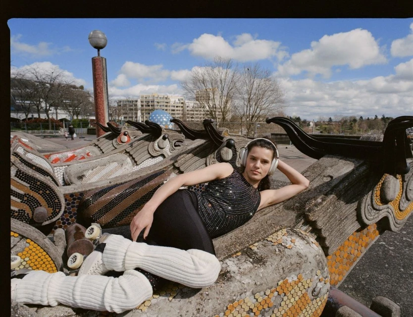 Woman with headphones relaxing on decorative dragon structure in a park setting on a sunny day with blue sky and clouds in the background.