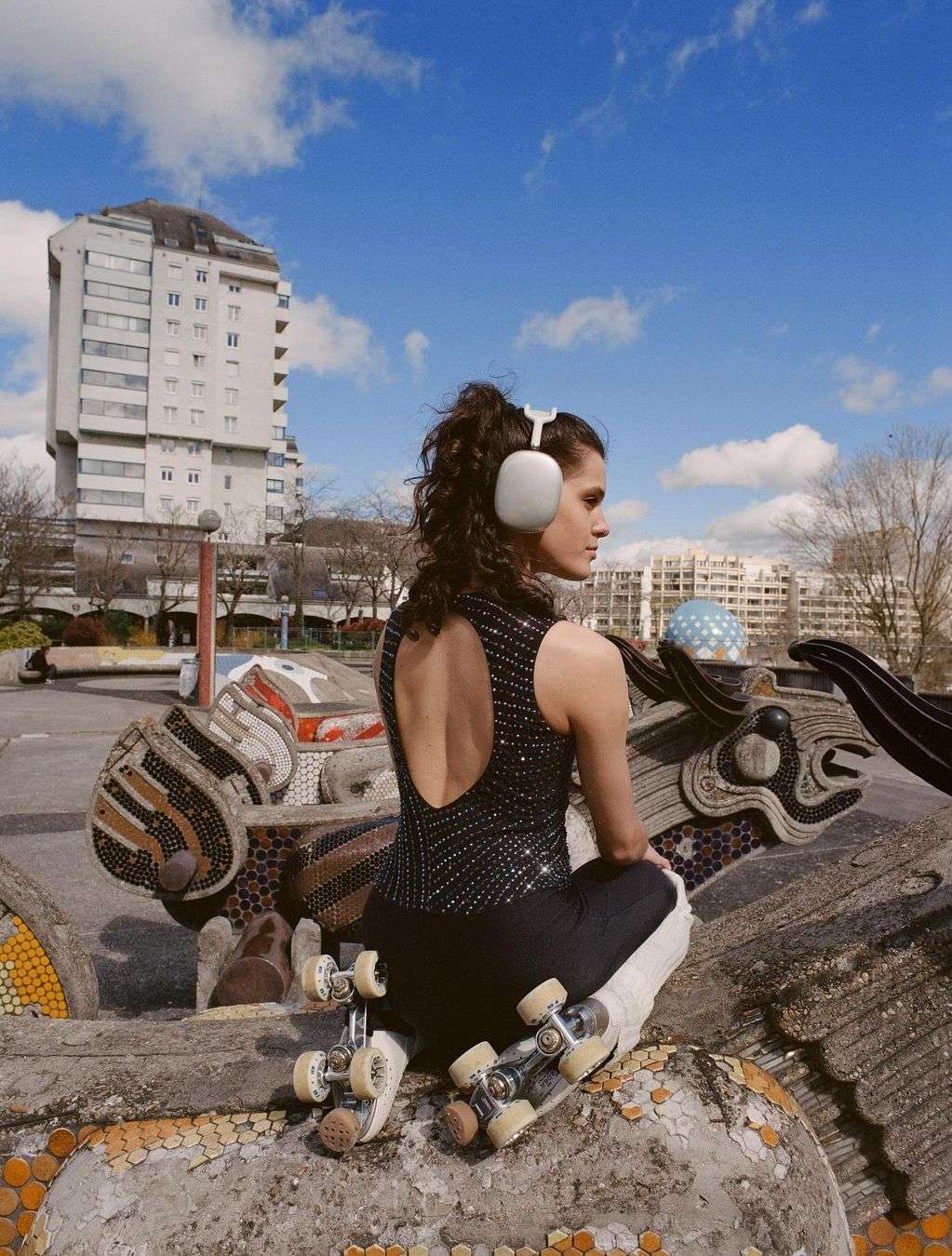 Young woman with curly hair and headphones poses with roller skates outdoors on a sunny day, featuring urban art and a modern building.
