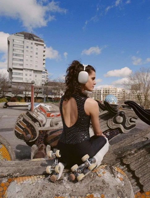 Young woman with curly hair and headphones poses with roller skates outdoors on a sunny day, featuring urban art and a modern building.