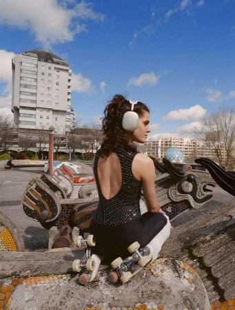 Young woman with curly hair and headphones poses with roller skates outdoors on a sunny day, featuring urban art and a modern building.