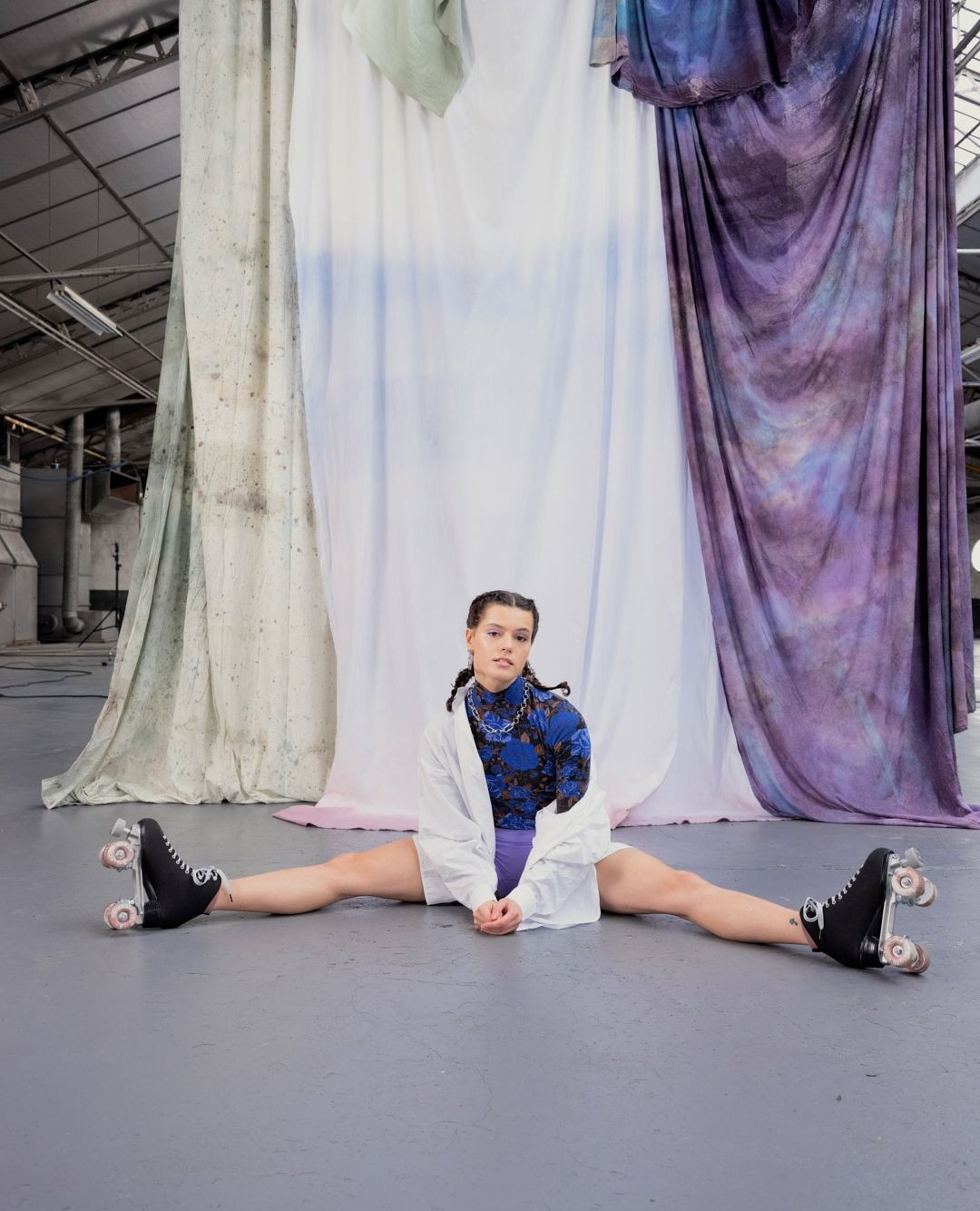 A young adult woman sits in a wide leg split on roller skates in a large studio space with textured fabric backdrops, and industrial ceilings.