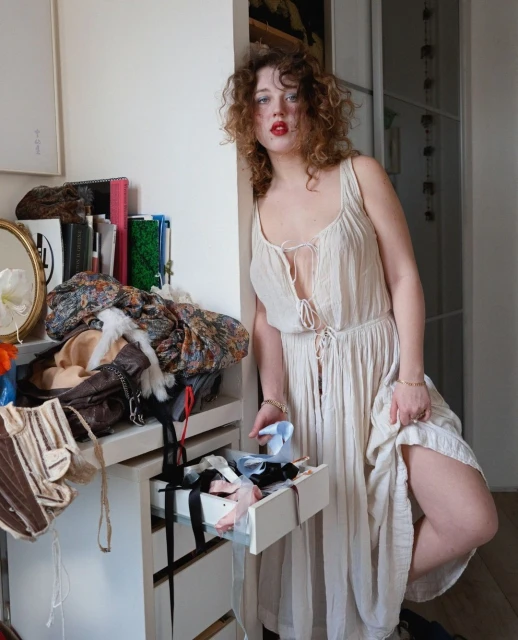 A woman with curly hair is leaning against a wall, wearing a cream-colored dress with a partially open drawer filled with clothes.