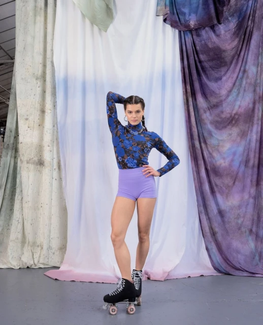 Full shot of a young adult woman in a floral top and shorts posing with retro roller skates in front of a patterned backdrop.