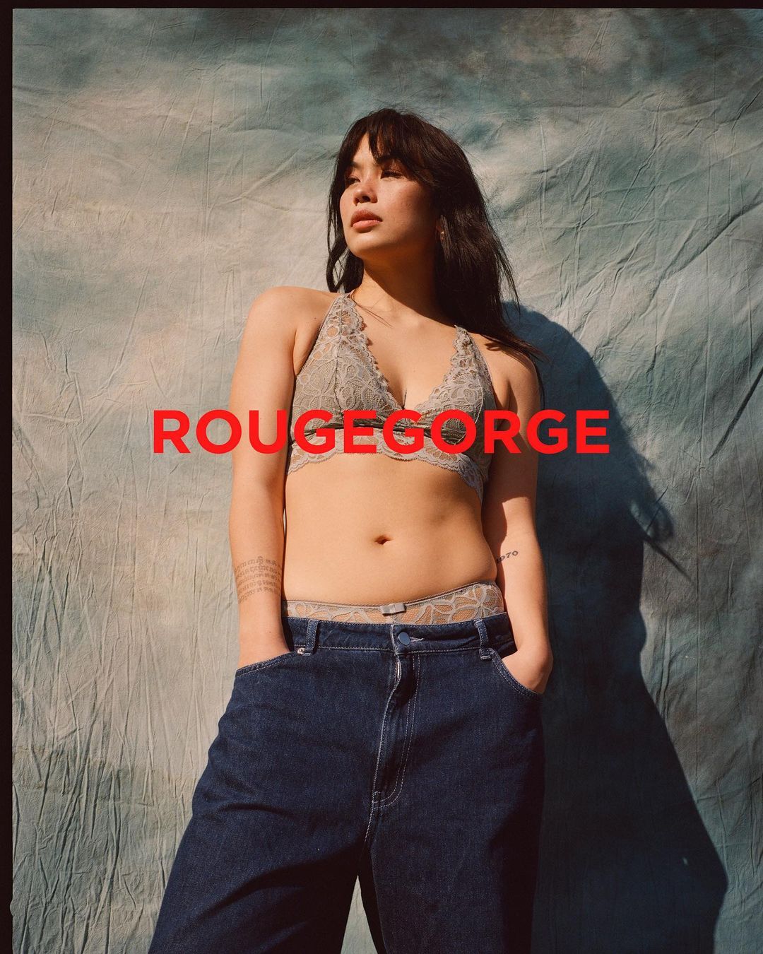 Woman in lace lingerie and oversized jeans posing against a mottled backdrop with the brand name 'RougeGorge' in red letters.