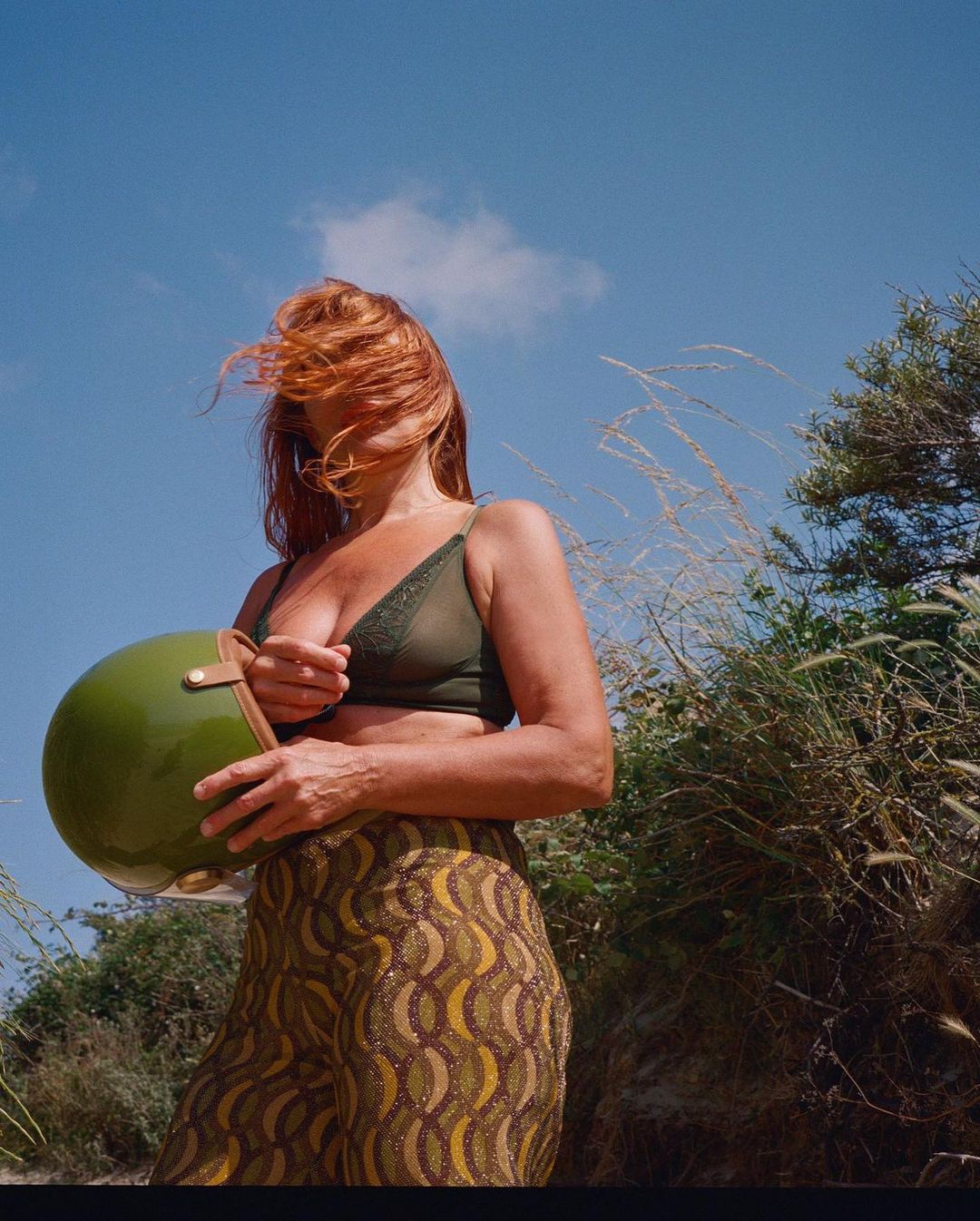 Woman with red hair holds a green motorcycle helmet outdoors on a sunny day, wearing a green top and patterned pants.
