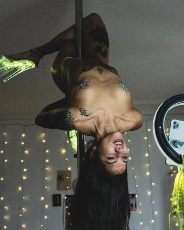 Adult woman performs a pole dance, inverted and upside down, inside a room with decorative string lights and illuminated ring light.