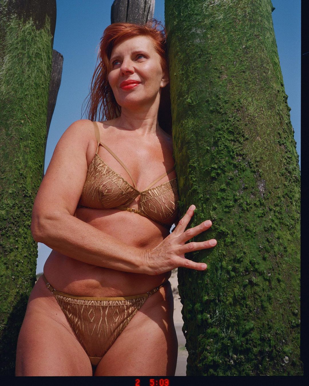 Red-haired woman in a golden bikini leans against moss-covered poles on a beach under a bright blue sky, showcasing beauty and swimwear fashion.