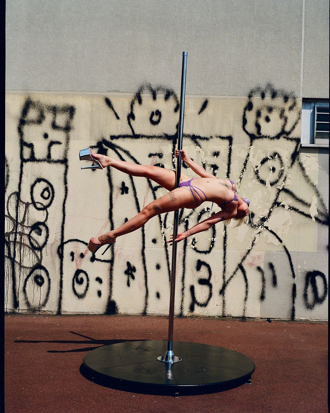 A female pole dancer in lilac lingerie gracefully poses outdoors with graffiti art behind her, showcasing strength and flexibility.