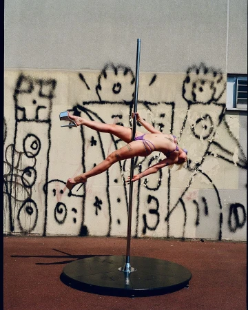 A female pole dancer in lilac lingerie gracefully poses outdoors with graffiti art behind her, showcasing strength and flexibility.