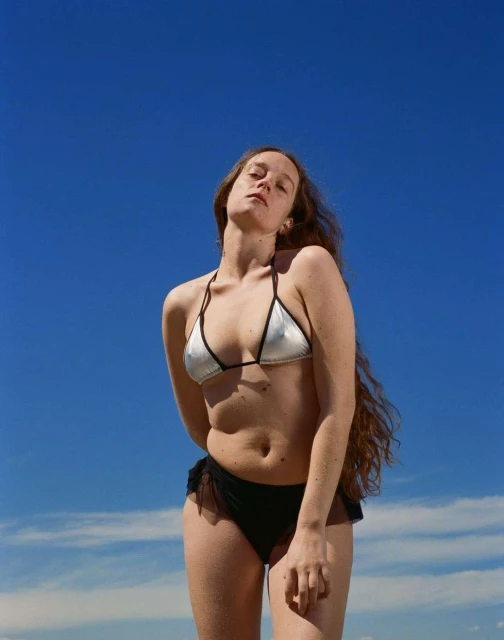 Low-angle shot of a young woman with long, wavy hair in a silver bikini top and black bottoms against a bright blue sky.