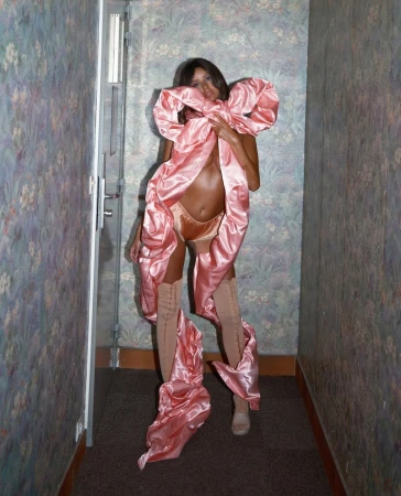 Woman in pink satin bow-dress with tan thigh-high boots poses in hallway with floral wallpaper and brown carpet, fashion concept.