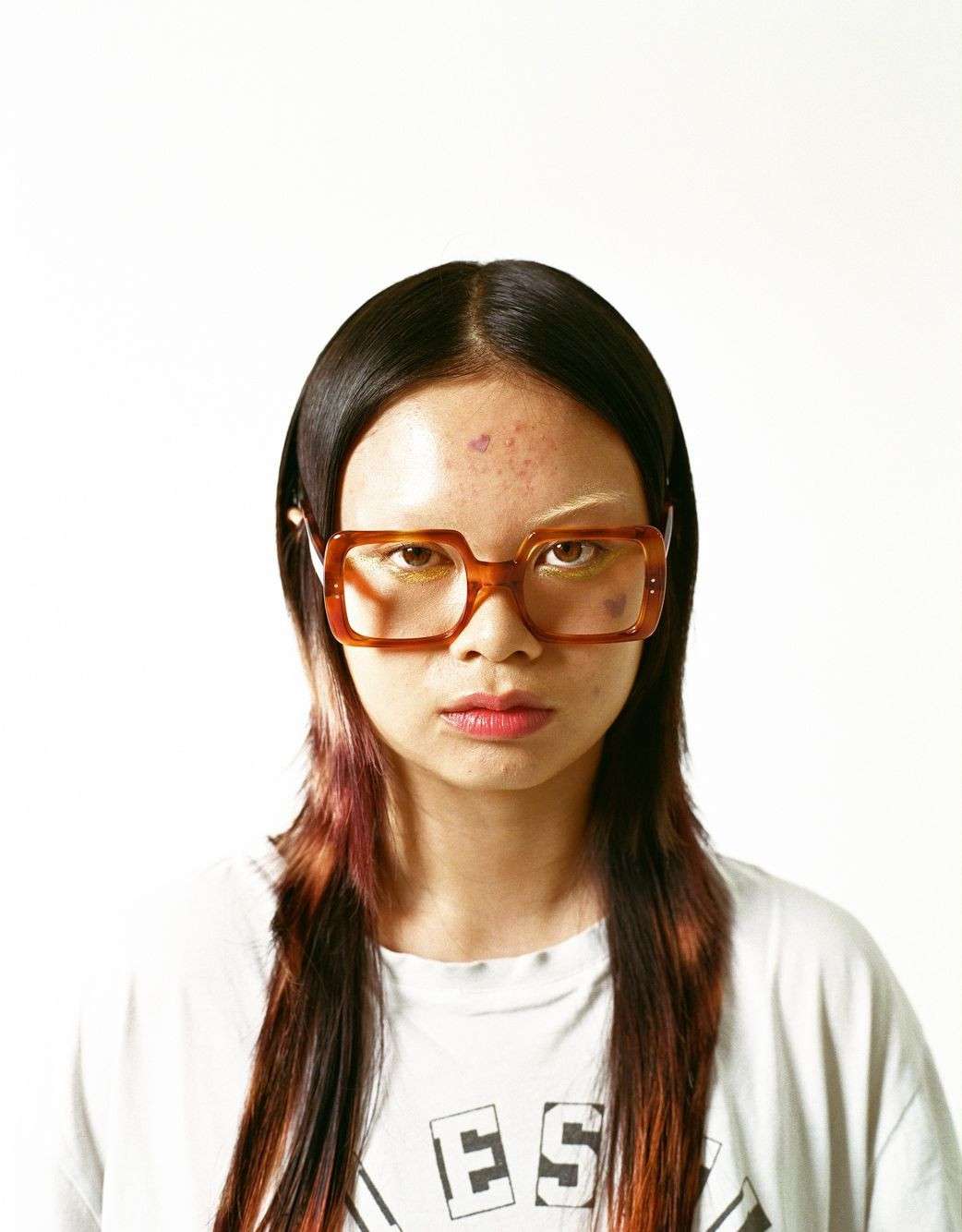 Portrait of a young adult woman with long dark hair and orange-framed glasses, wearing a white printed t-shirt against a white background.