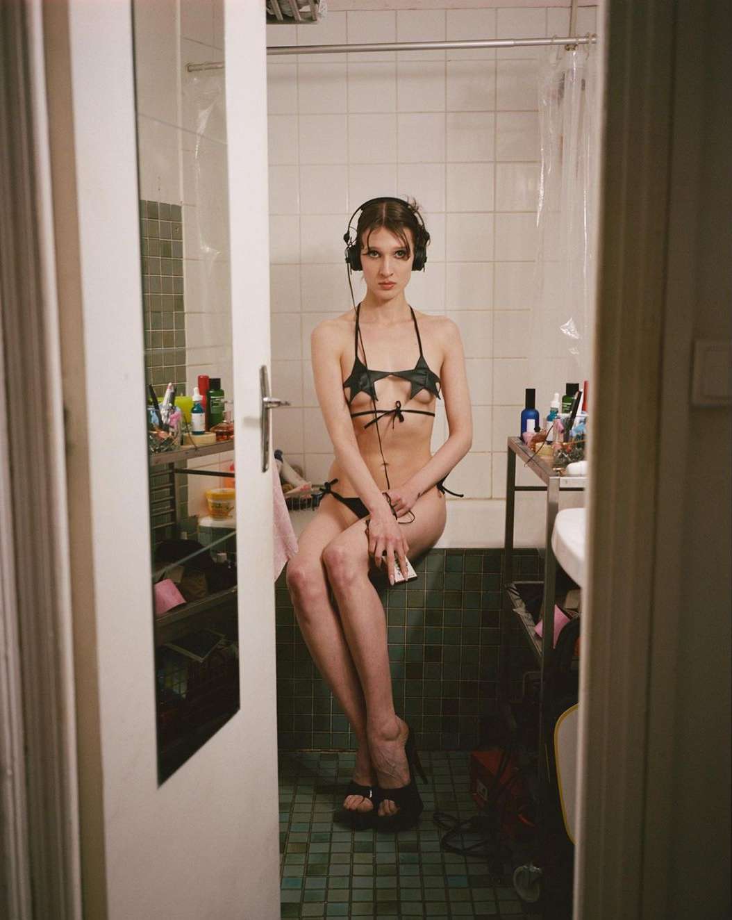 A young woman with headphones in a bikini sits on the edge of a bathtub in a brightly lit bathroom with an open door in the foreground.
