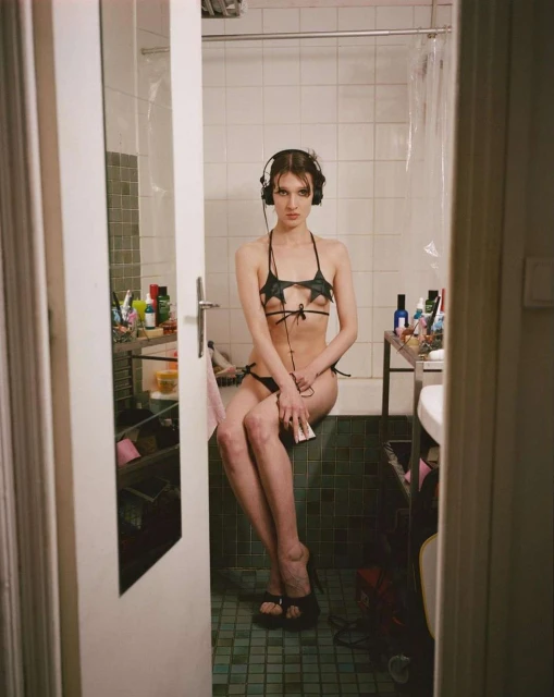 A young woman with headphones in a bikini sits on the edge of a bathtub in a brightly lit bathroom with an open door in the foreground.