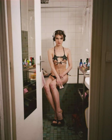 A young woman with headphones in a bikini sits on the edge of a bathtub in a brightly lit bathroom with an open door in the foreground.