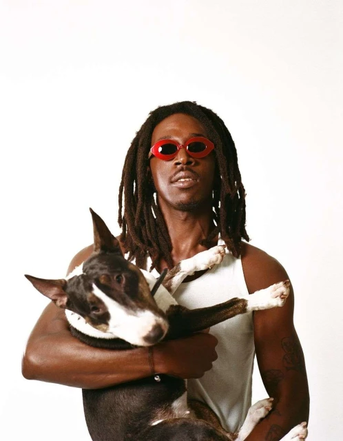 A man with dark skin and dreadlocks is holding a bull terrier dog while wearing red sunglasses against a bright white backdrop.