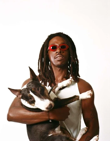A man with dark skin and dreadlocks is holding a bull terrier dog while wearing red sunglasses against a bright white backdrop.