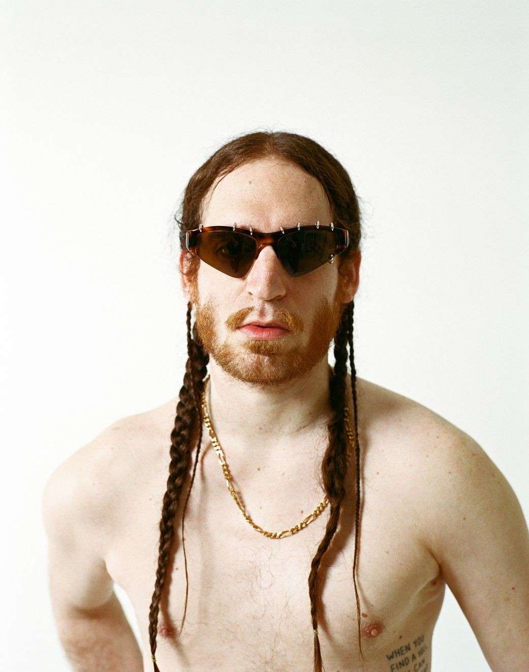 Portrait of a shirtless adult male with reddish hair in braids, wearing sunglasses and a gold chain, against a white background.
