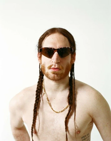 Shirtless man with red braided hair, tortoiseshell sunglasses, and a gold chain against a white background by Camille Mompach