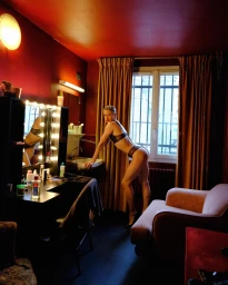 Woman in black lingerie standing near a lit vanity mirror and window in a red hotel room by Camille Mompach