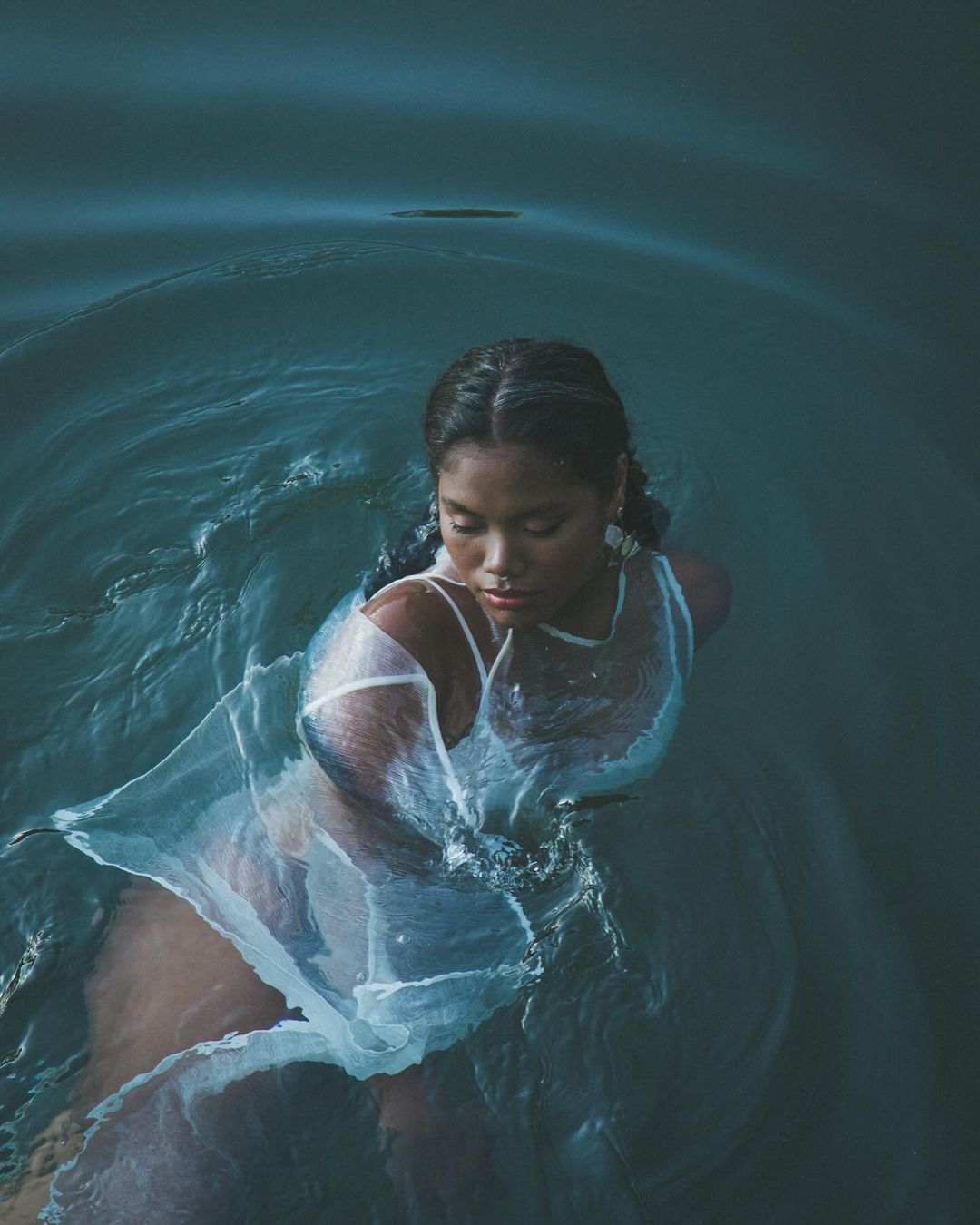 Young adult female in translucent dress, waist-deep in calm water with ripples and dark blue tones, seen from an eye-level, high-angle view.