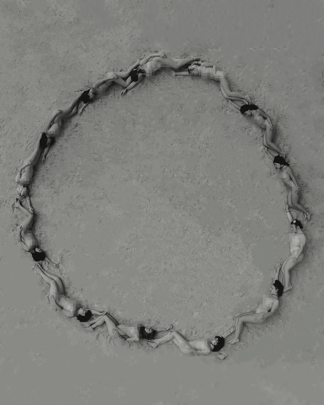 Aerial shot displays multiple nude adults in a circular pattern on gray sand-like surface, creating unique artistic human formation.