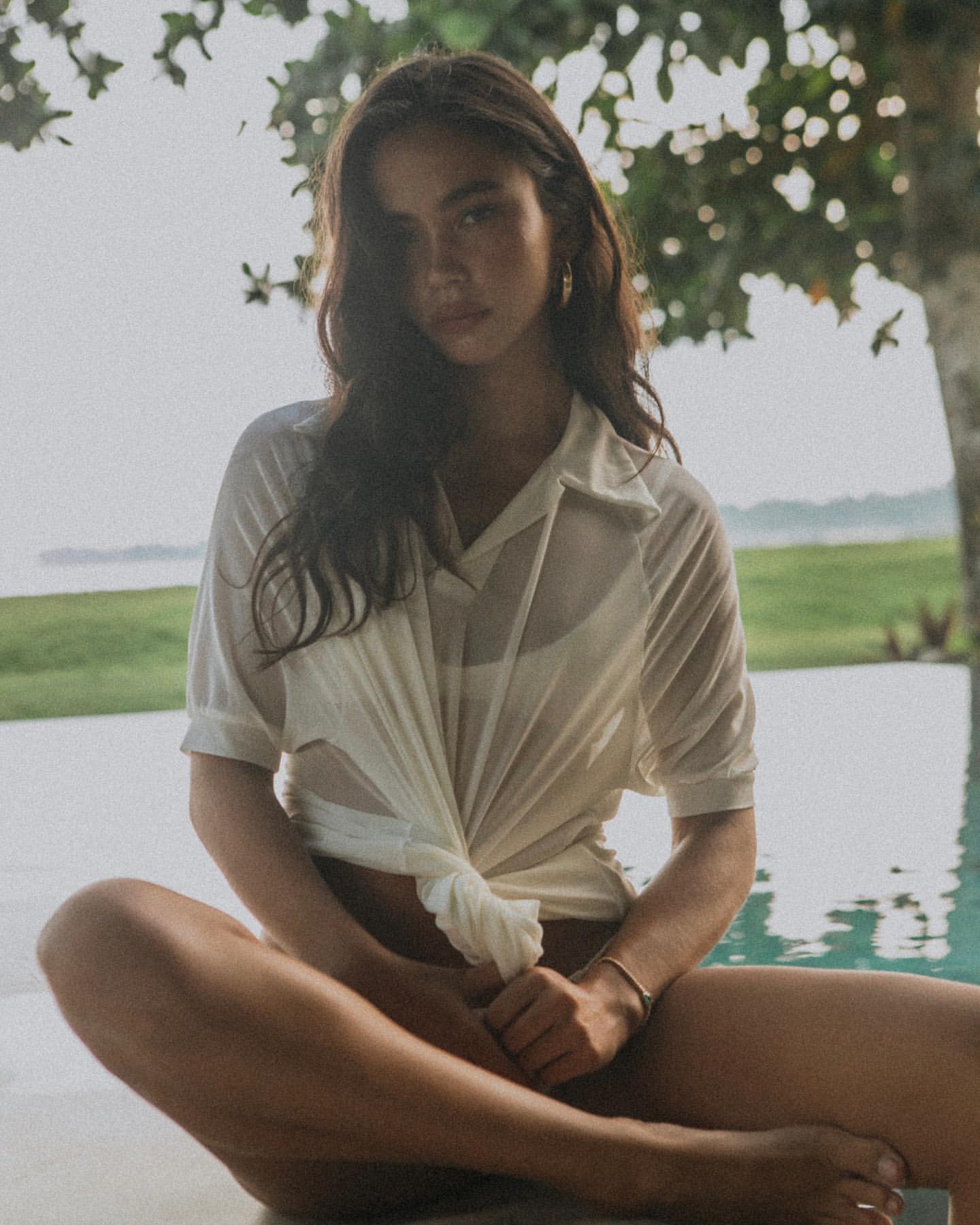 Adult female with dark hair and tan skin sitting in front of swimming pool wearing a white shirt and bottoms outdoors during daytime.