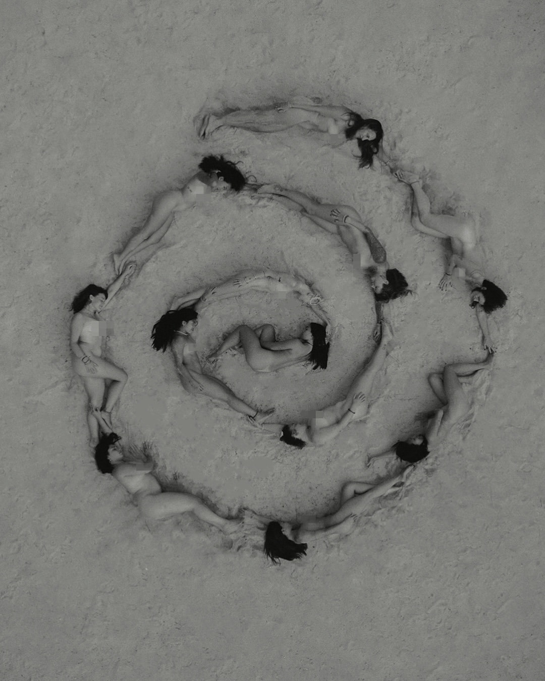 Overhead shot of a spiral formation of nude adult women lying on a grainy surface, forming a circular shape in a monochromatic tone.