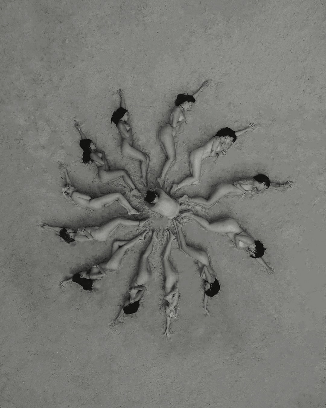 Aerial shot of several women lying in a circle formation on a sandy surface in monochrome, creating a visually striking composition.