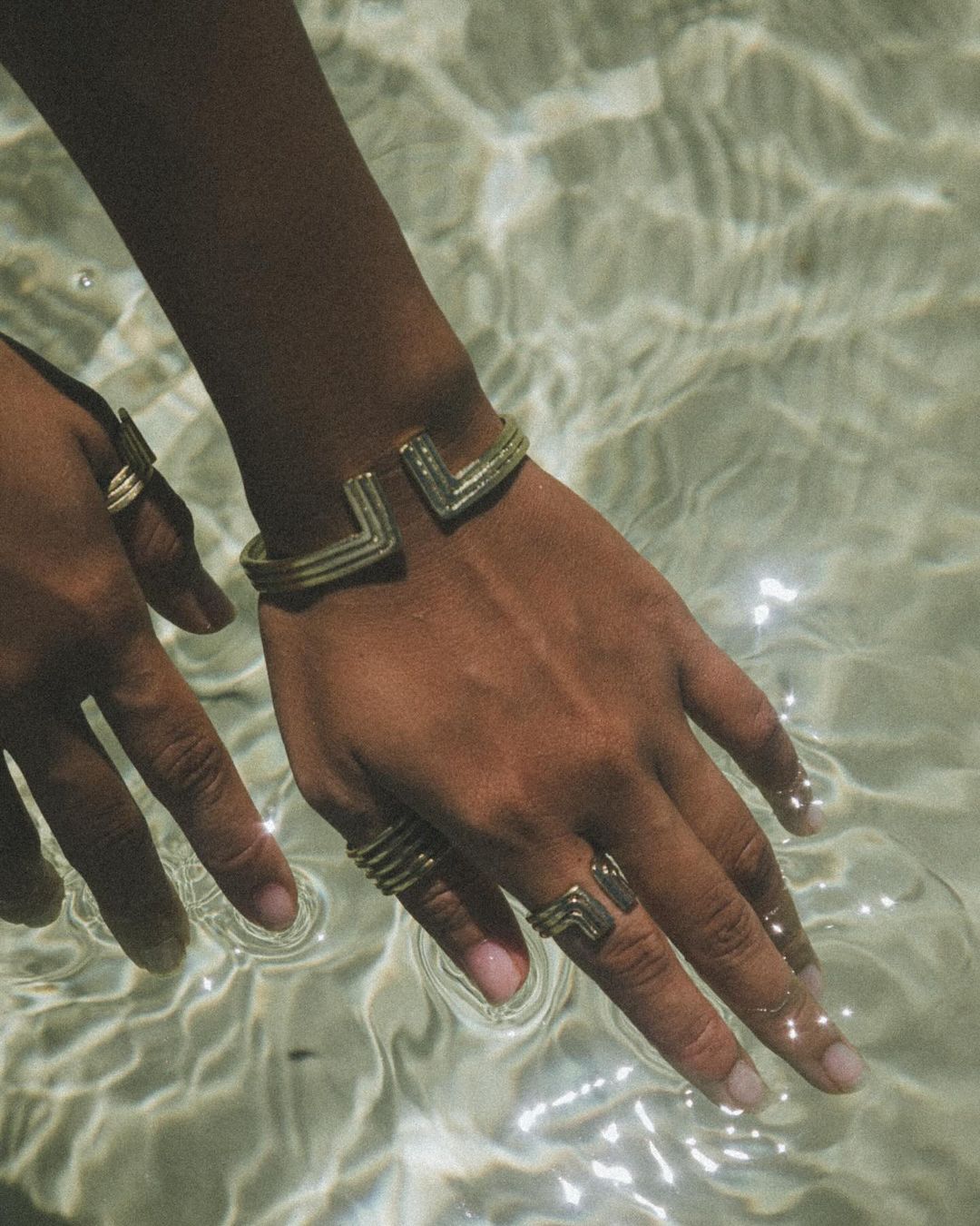 Close-up shot of an arm and hand with tan skin wearing several decorative gold rings and a gold cuff bracelet near water.