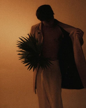 Man wearing an unbuttoned coat holding a green palm frond against a warm-toned background, photographed by Camille Robiou Du Pont