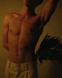 Shirtless man wearing light trousers holding a dark frond plant, photographed by Camille Robiou Du Pont