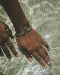 Close-up of hands wearing gold tone metal bangles and rings submerged in shallow rippling water