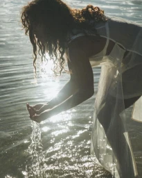 Woman in a sheer white dress bending over rippling water, letting water pour from her cupped hands with bright sunlight gleaming