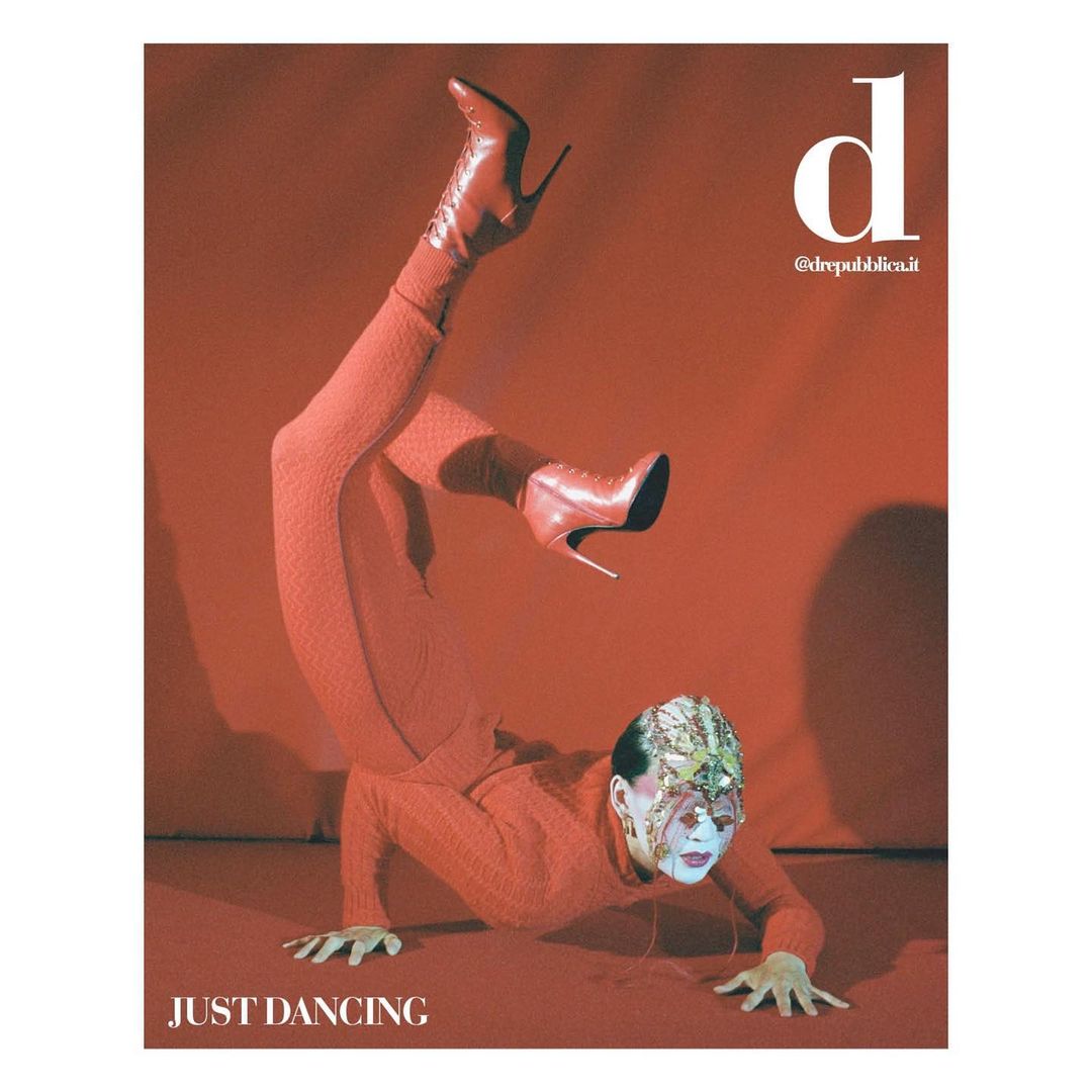 A person in red clothing and high-heeled boots performing a handstand on a red background, appearing on a magazine cover with white text.