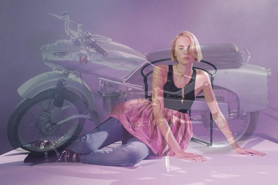 Woman posing next to a vintage motorcycle with a muted purple background. The woman is lying down on a white surface.