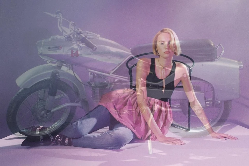 Woman posing next to a vintage motorcycle with a muted purple background. The woman is lying down on a white surface.