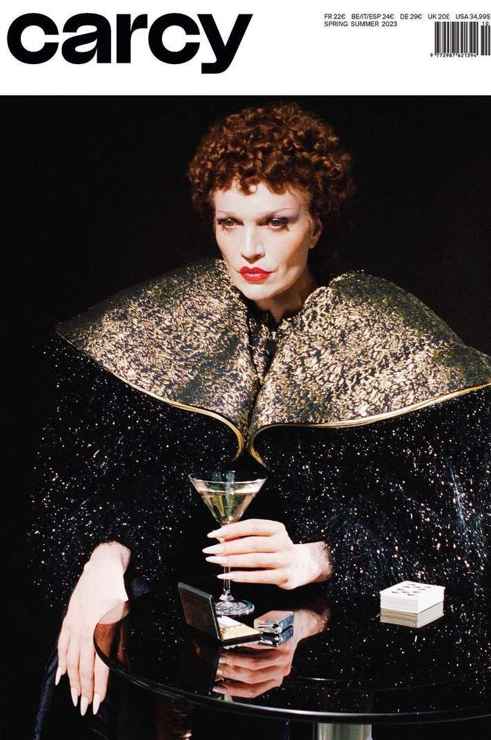 Model with short curly brown hair and red lipstick wearing a heavily textured black and gold collared jacket holding a martini glass at a table