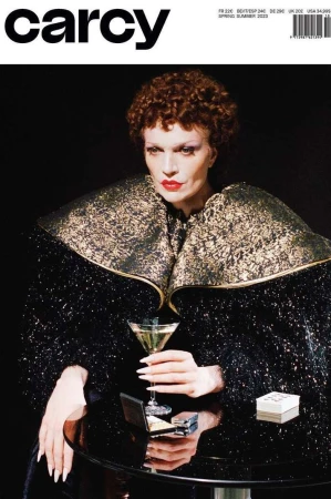 Model with short curly brown hair and red lipstick wearing a heavily textured black and gold collared jacket holding a martini glass at a table