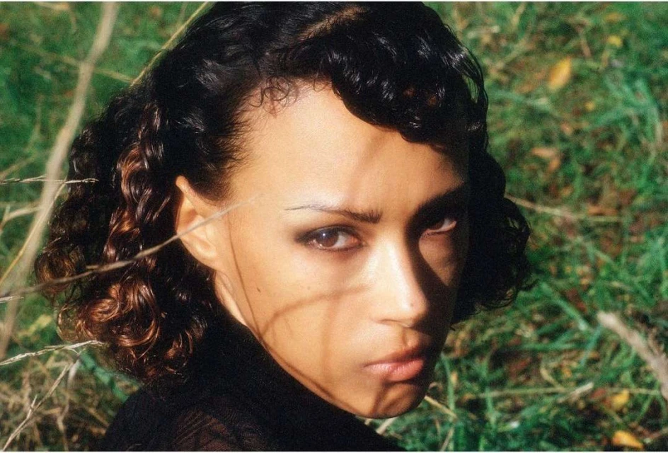 Portrait of an adult woman with dark curly hair and brown eyes, staring seriously into the camera in a grassy outdoor setting.