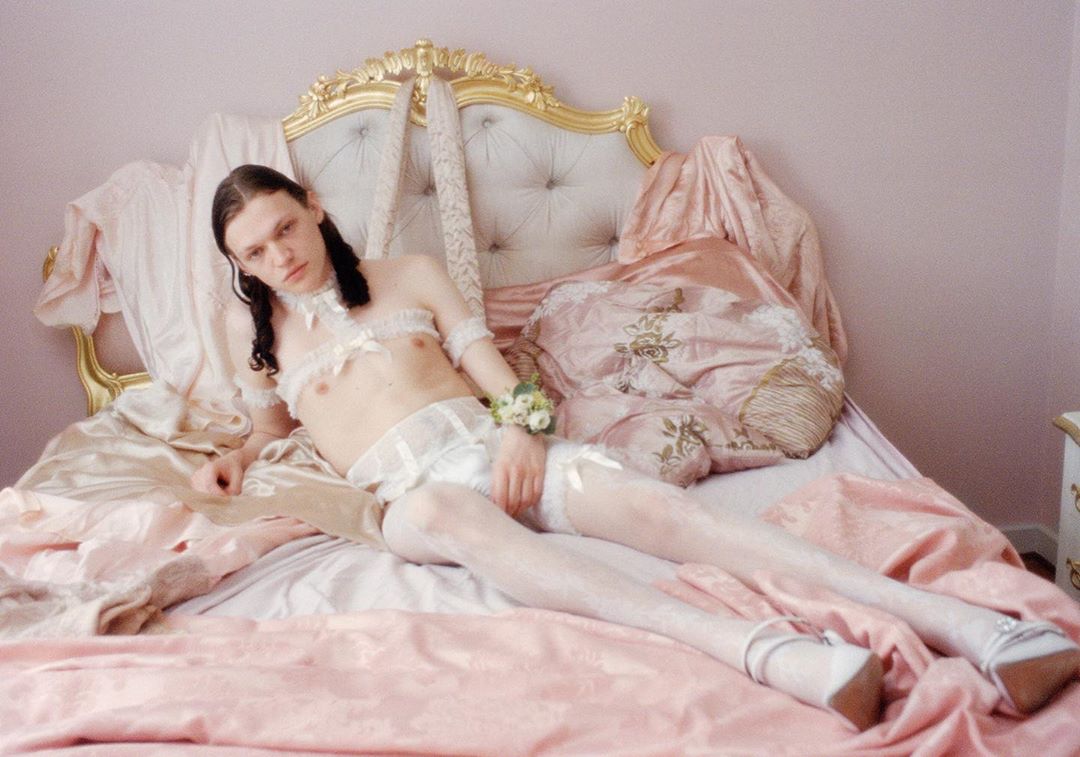 Non-binary person in white lingerie and silver heels reclines on a pink and gold bed, looking at the viewer with a neutral expression.