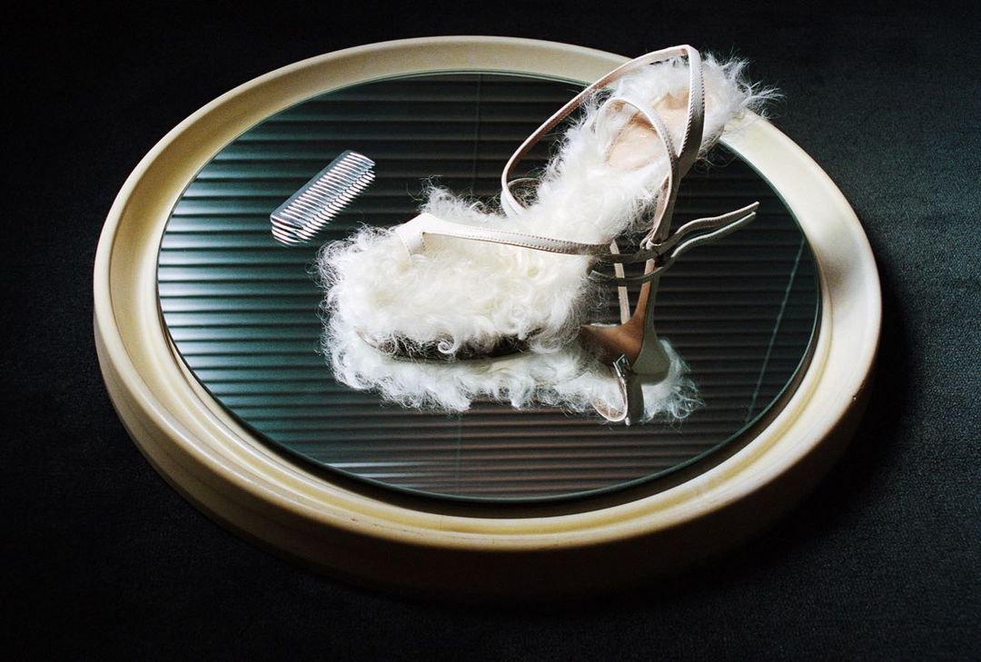 High heel sandal with fluffy white fur trim displayed on a round mirrored surface with black louvers and a silver comb accent.