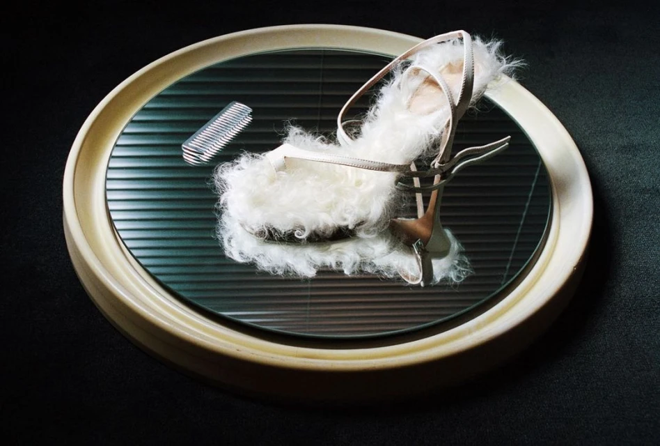 High heel sandal with fluffy white fur trim displayed on a round mirrored surface with black louvers and a silver comb accent.