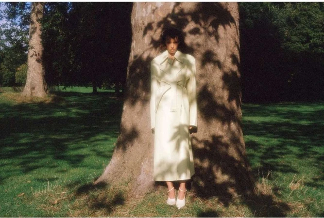 Model wearing a light yellow coat leaning against a large tree trunk in a park setting.