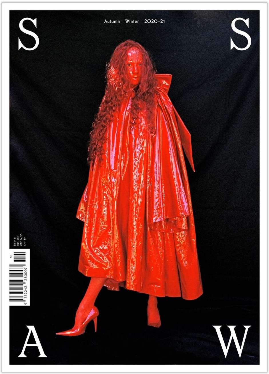 Person in red vinyl outfit and high heels poses against dark background for magazine cover with white 'S' and 'A' and 'W' letters.