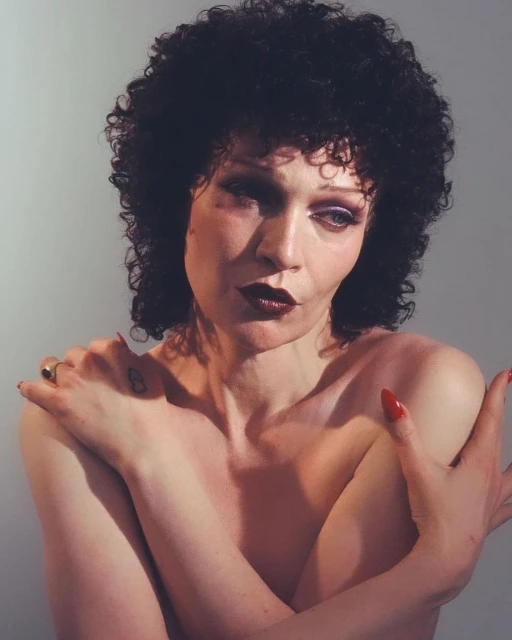 Striking close-up portrait of a non-binary person with dark curly hair and bold makeup, posing with arms crossed in a neutral studio setting.